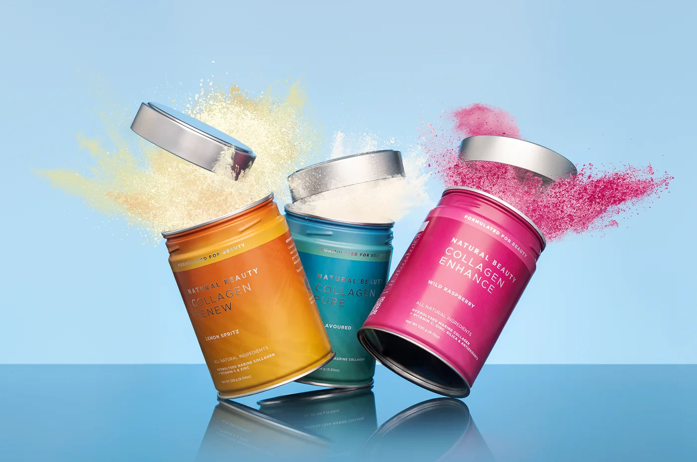 Three Natural Beauty collagen tins bursting open with lemon, white and raspberry powder on a pale blue background