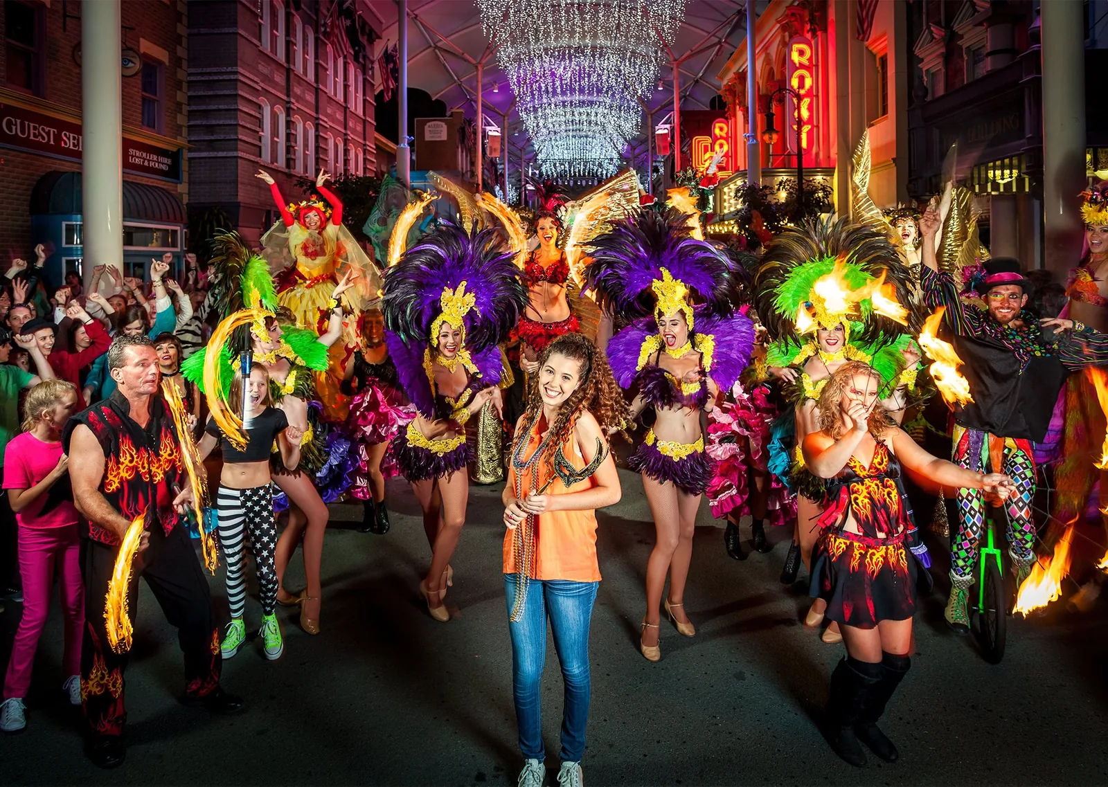 A night street parade at Movie World: a smiling girl in front of feathered dancers, fire twirlers and cheering crowds under neon signs