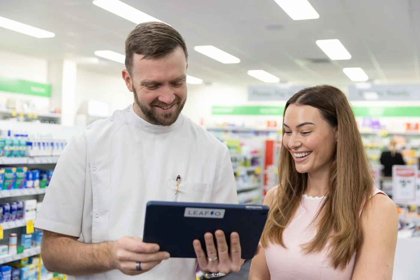 A smiling pharmacist showing a customer a Leafio tablet in a pharmacy aisle